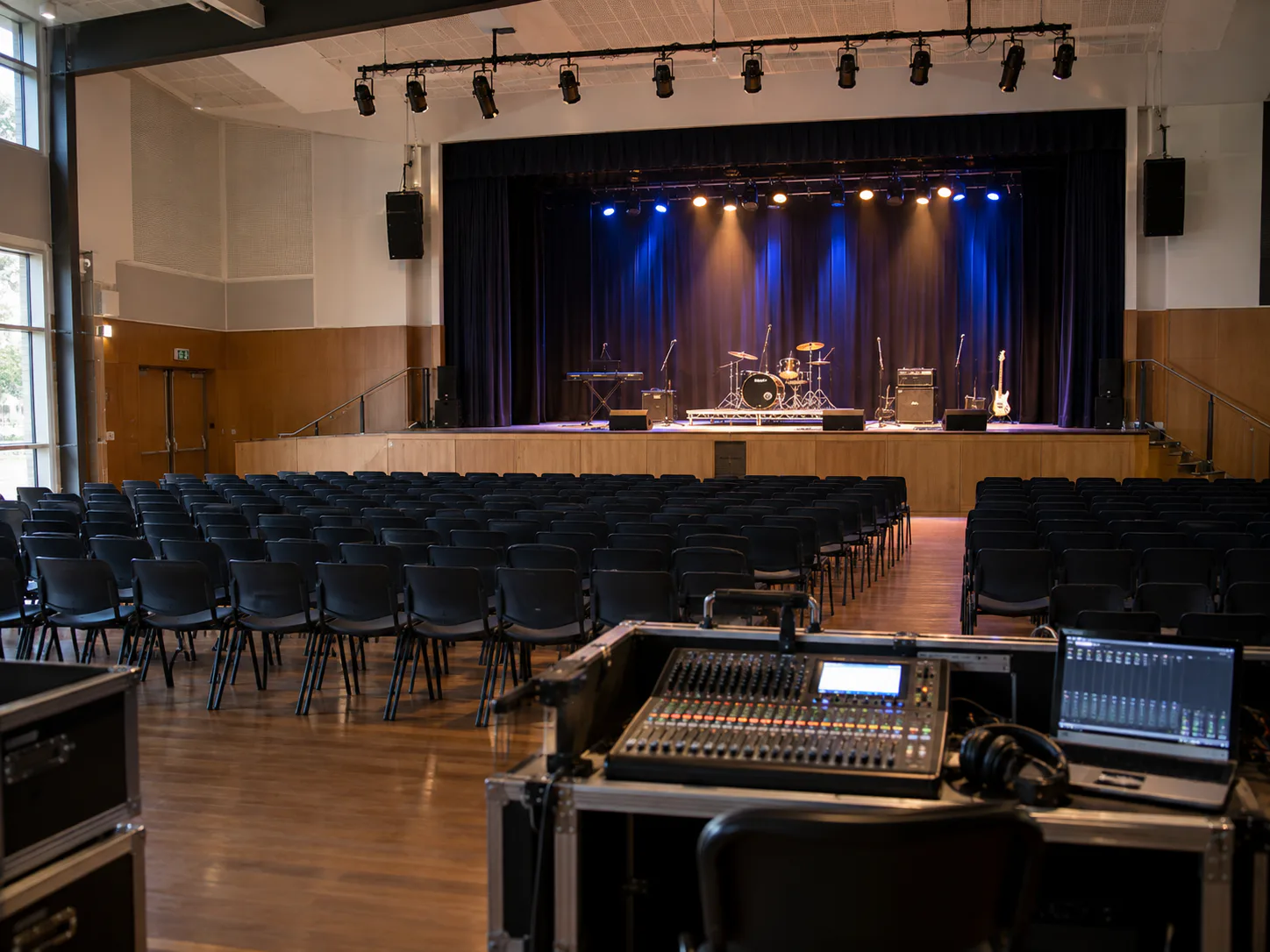 School hall from the operator position — digital mixing console and laptop in the foreground, stage lighting and speakers beyond