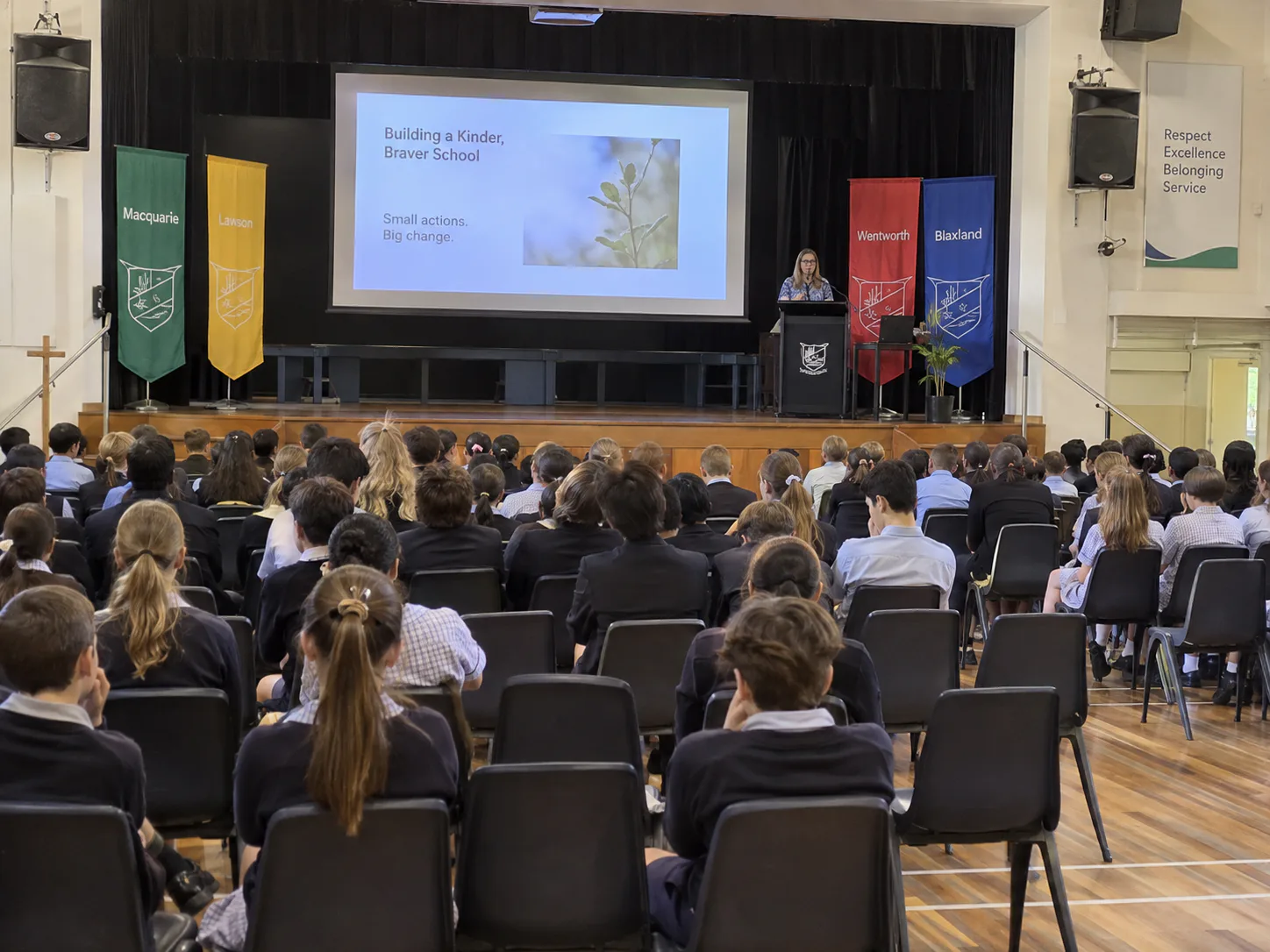 Students seated in a school hall during assembly, presenter at a lectern with a projection screen