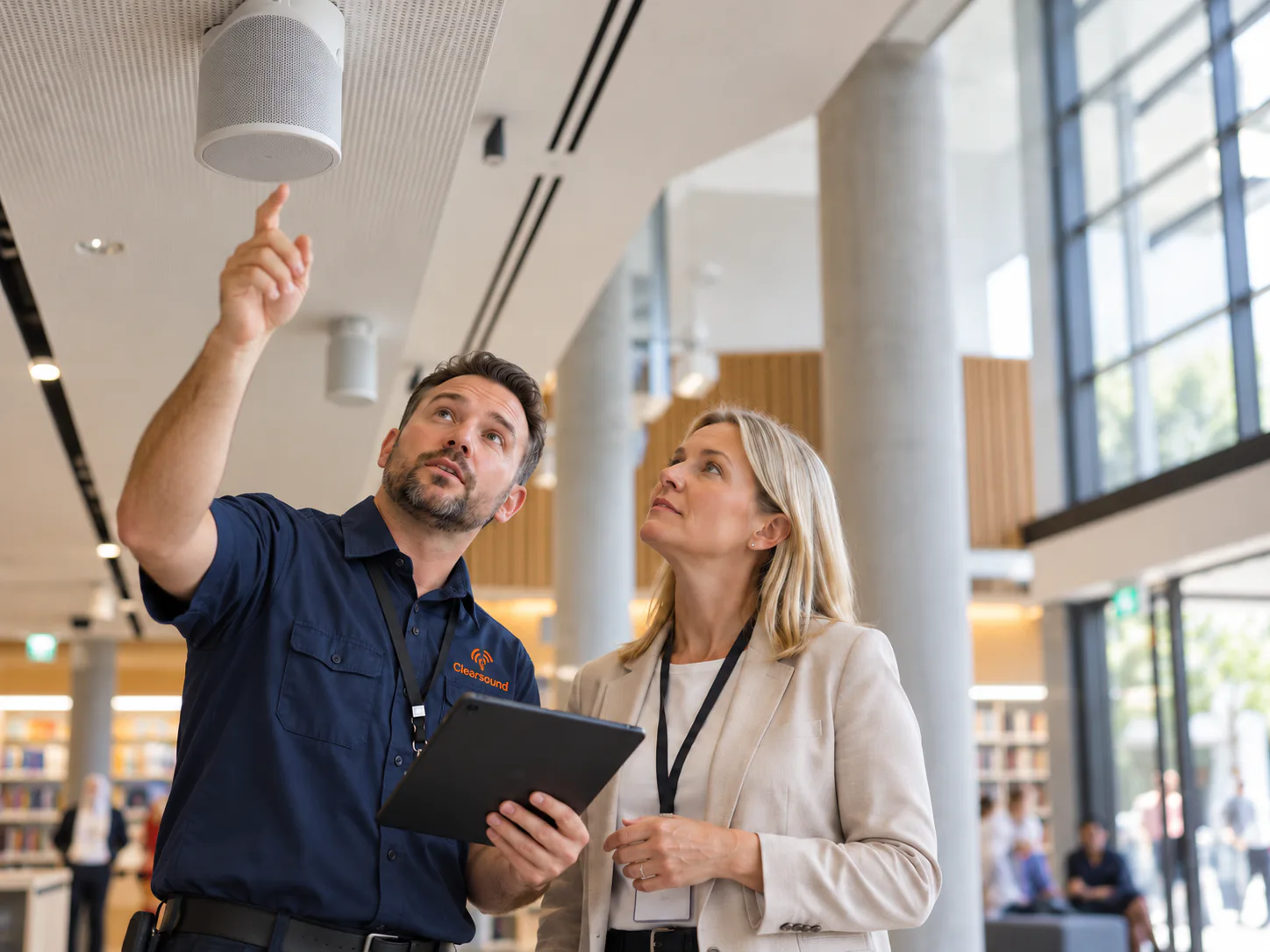 Technician pointing to a ceiling speaker in a library foyer while a manager looks on