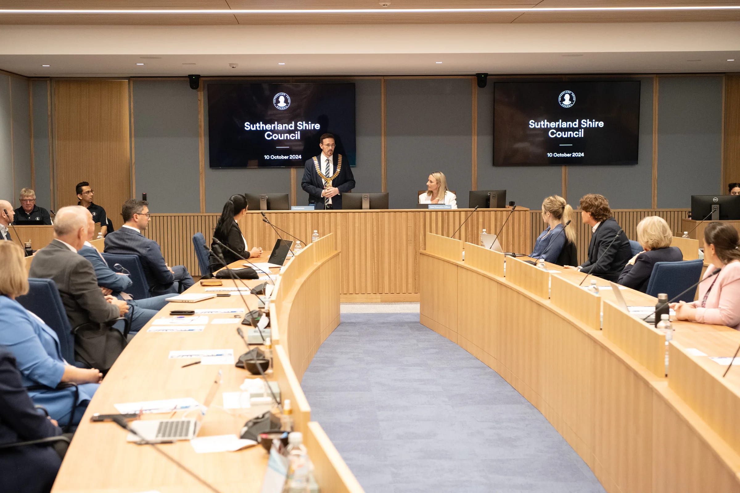 Council chamber in session, mayor addressing councillors seated at curved desks with microphones and displays